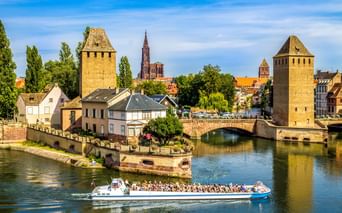 Touristenboot auf Fluss vor mittelalterlichen Türmen und Brücke in Straßburger Altstadt, mit Münsterspitze im Hintergrund unter blauem Himmel.