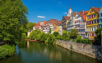 Bunte Fachwerkhäuser mit roten Dächern säumen den Neckar in Tübingens Altstadt. Ein Kirchturm ragt über den Gebäuden unter blauem Himmel empor.