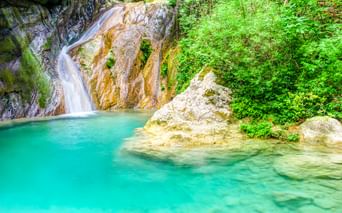 Wasserfall, der über goldbraune Felsen in ein türkisfarbenes Becken bei Nydri, Lefkada fließt. Üppige grüne Vegetation umgibt das natürliche Becken.