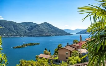Panoramablick auf den Lago Maggiore mit kleinen Inseln, umgeben von Bergen. Dorfhäuser mit roten Dächern und Kirchturm im Vordergrund, Palmwedel sichtbar.