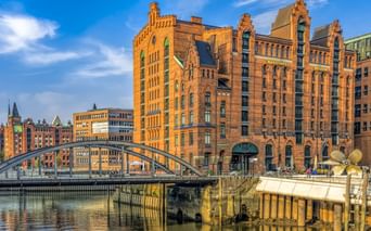 Rote Backsteingebäude der Hamburger Speicherstadt mit Bogenbrücke über Kanal. Historische Gebäude spiegeln sich im Wasser unter blauem Himmel.