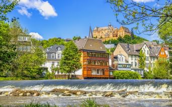 Blick auf Marburg mit Flusswehr im Vordergrund, historischen Fachwerkhäusern am Flussufer und dem Marburger Schloss auf dem Hügel.