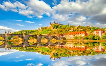 Historic stone bridge with multiple arches spanning the Main River in Würzburg, reflecting in calm water. Marienberg Fortress crowns the hilltop above.