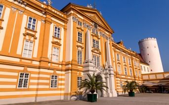 Barocke Fassade von Stift Melk in Gelb und Weiß mit prächtigem Eingangsportal. Palmen im Innenhof, weißer Turm rechts sichtbar.