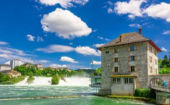 Rheinfall bei Schaffhausen mit kraftvoll strömendem Wasser. Historisches Steingebäude steht neben dem türkisfarbenen Fluss unter blauem Himmel.