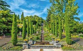 Formale Terrassengärten auf der Insel Mainau mit Steinbrunnen, Zypressen, bunten Blumenbeeten und breiten Steintreppen unter blauem Himmel.