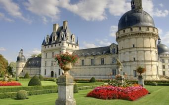 Französisches Renaissance-Schloss mit runden Türmen und Kuppeldächern. Gepflegte Gärten mit roten Blumen, geschnittenen Hecken und Steinurnen im Vordergrund.