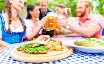 Bayerische Brotzeit auf einem Holzbrett mit Obatzda im Vordergrund. Menschen in Dirndl und Freizeitkleidung stoßen mit Bierkrügen an.