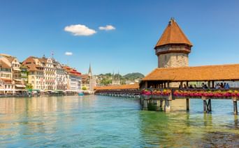 Historische Kapellbrücke mit orangem Dach und achteckigem Wasserturm über der Reuss in Luzern, mit bunten Gebäuden entlang des Ufers.