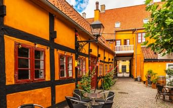 Enge Kopfsteinpflastergasse in Køge mit leuchtend orangen Fachwerkhäusern, rotgerahmten Fenstern, orangen Ziegeldächern und Café-Bestuhlung.