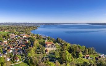Luftaufnahme von Bernried am Starnberger See mit Dorf am Seeufer, rotgedeckten Häusern, großem historischen Gebäude und blauem Seewasser unter klarem Himmel.