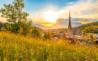 Wallfahrtskirche Maria Alm mit hohem Turm im österreichischen Dorf bei Sonnenuntergang, umgeben von Wildblumenwiese und Bergen unter goldenem Himmel.
