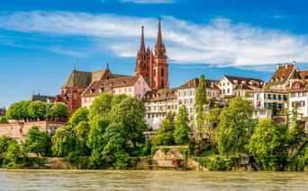 Blick auf die Basler Altstadt vom Rhein mit den beiden Türmen des roten Sandstein-Münsters über historischen Gebäuden und grünen Bäumen.