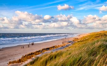 Breiter Sandstrand in Julianadorp mit grasbewachsenen Dünen im Vordergrund, Spaziergängern am Ufer und weißen Wolken über der Nordsee.