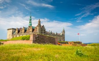 Schloss Kronborg in Helsingør mit grünen Kupfertürmen und Steinbefestigungen, umgeben von Grashängen. Dänische Flagge weht über den Mauern.