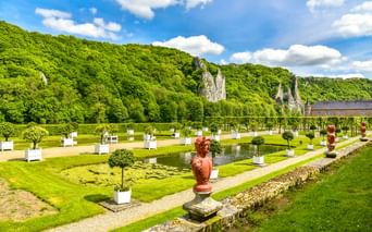 Formaler Garten in Freyr mit Zierteich, geschnittenen Bäumen, roten Statuen und dramatischen Kalksteinfelsen im Hintergrund unter blauem Himmel.
