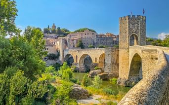 Mittelalterliche Steinbrücke mit Turm und Bögen über einem Fluss in Besalú, Spanien. Historische Gebäude und grüne Vegetation umgeben die Brücke.