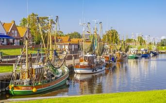 Traditionelle Fischerboote im Hafen von Greetsiel mit Häusern mit gelben Dächern entlang des Ufers und grünen Grasflächen unter blauem Himmel.