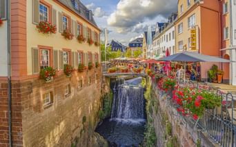 Wasserfall im historischen Zentrum von Saarburg, flankiert von bunten Gebäuden mit Blumenkästen und Straßencafés unter Sonnenschirmen.