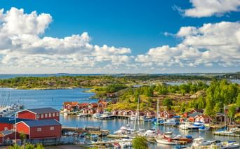 Hafen im Göteborger Schärengarten mit traditionellen roten Holzhäusern und Booten. Felsige Inseln mit grüner Vegetation unter blauem Himmel.