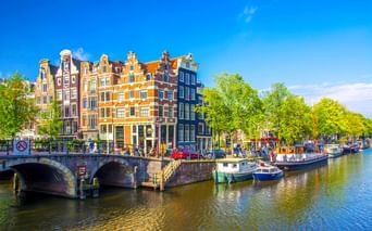 Amsterdamer Gracht mit traditionellen niederländischen Giebelhäusern in verschiedenen Farben, Booten am Ufer und Steinbrücke unter blauem Himmel.
