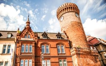 Historischer Spremberger Turm in Cottbus mit Zifferblatt und Zinnen, neben rotem Backsteingebäude mit verzierten Giebeln unter blauem Himmel.