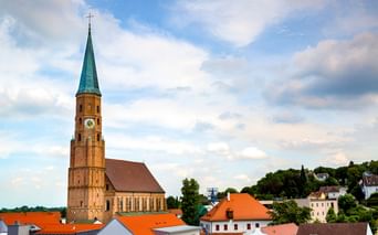 Spätgotische Hallenkirche St. Johannes in Dingolfing mit hohem Backsteinturm, grüner Spitze und Uhr, überragt rotgedeckte Häuser.