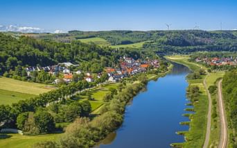 Aerial view of Beverungen with red-roofed houses along the Weser River, surrounded by green forests and hills. Wind turbines visible on distant hills.