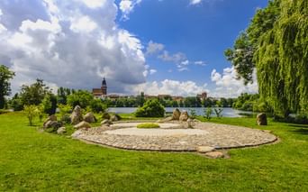 Kreisförmiger Steingarten mit großen Findlingen und Kies auf grüner Wiese am Tiefwarensee in Waren-Müritz. Kirchturm über dem See sichtbar.