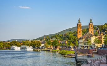 View of Miltenberg with St. Jakobus Church's twin towers along the Main River. Riverfront promenade with boats, hills in background.