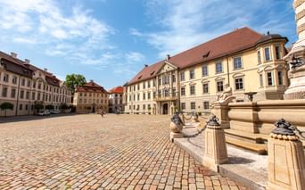 Kopfsteinpflasterplatz in Eichstätt mit verziertem Brunnen und gelben Barockpalais mit roten Ziegeldächern unter blauem Himmel.