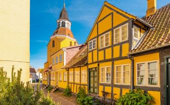 Gelbe Fachwerkhäuser mit Kirchenglockenturm in Faaborg, Dänemark. Die historischen Gebäude zeigen traditionelle Architektur unter blauem Himmel.