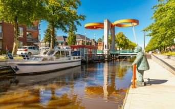 Bunte Eurobrug-Zugbrücke mit gelb-roten Scheiben über Kanal in Stadskanaal. Boote am Ufer, grüne Bäume und Statue sichtbar.