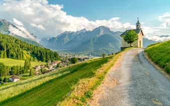 Kleine Kapelle mit Glockenturm an einem Schotterweg mit Blick auf Toblach und die Dolomiten unter blauem Himmel mit weißen Wolken.
