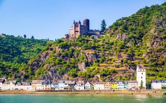 View of St. Goar on the Rhine with medieval castle on terraced vineyard hillside. Colorful buildings line the riverbank below steep slopes.
