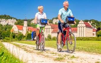 Two cyclists on red bikes riding on a gravel path with Beuron Monastery and limestone cliffs in the background under clear blue sky.