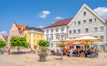 Sonniger Marktplatz in Schongaus Altstadt mit bunten historischen Gebäuden, Straßencafé mit Sonnenschirmen, Brunnen und Menschen.