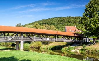 Überdachte Holzbrücke mit rotem Dach über einem ruhigen Fluss in Fridingen. Grüne Wiese im Vordergrund, bewaldete Hügel und Häuser im Hintergrund.