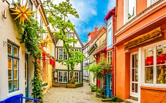 Schmale Kopfsteinpflasterstraße in Bremen, gesäumt von bunten Fachwerkhäusern in Gelb, Orange und Rot. Grüne Pflanzen und Sterne schmücken die Fassaden.