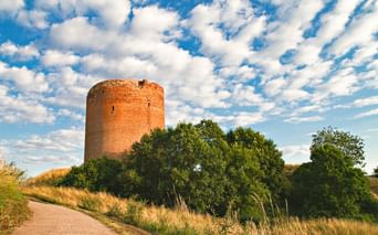 Round brick tower on a hill surrounded by green trees and grass under a blue sky with white clouds. A path leads toward the structure.