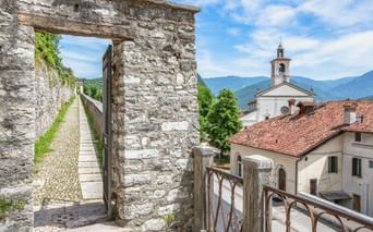 Alter Steinbogen mit Holztor entlang eines schmalen Weges in Feltre, mit weißem Kirchturm und Bergen im Hintergrund.