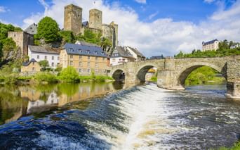 Mittelalterliche Burg auf einem Hügel über einer Steinbogenbrücke, die einen Fluss mit Wehr in Runkel überspannt. Bunte Häuser säumen das Flussufer.