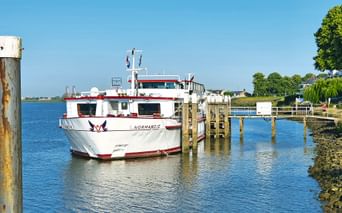 Weiß-rote Passagierfähre Normandie an einem Holzsteg in ruhigem blauen Wasser unter klarem Himmel, mit grünen Bäumen am Ufer.