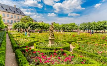 Barocker Rosengarten mit geschnittenen Hecken und bunten Rosen an der Neuen Residenz Bamberg. Steinstatue in der Mitte, historisches Gebäude dahinter.