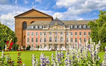Kurfürstliches Palais mit rosa Rokoko-Fassade und Konstantinbasilika in Trier. Bunte Blumenbeete blühen im Palastgarten unter blauem Himmel.
