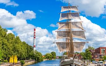 Großes Segelschiff mit weißen Segeln am Dane-Fluss in Klaipeda, Litauen. Grüne Bäume säumen den Wasserweg unter blauem Himmel mit Wolken.