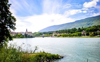 View across the Rhône River in Seyssel, France, showing a church tower and historic buildings along the waterfront with green mountains in the background.