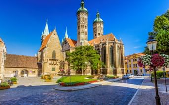 Naumburger Dom mit zwei hohen Türmen und orangefarbenen Dächern auf einem sonnigen Platz. Blumenbeete und Bäume schmücken den Kopfsteinpflasterplatz.