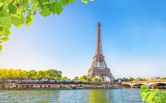Eiffelturm über der Seine in Paris an einem sonnigen Tag, eingerahmt von grünen Blättern. Eine Brücke und baumgesäumtes Flussufer sind sichtbar.