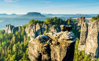 Blick von der Bastei in der Sächsischen Schweiz mit dramatischen Sandsteinsäulen über grünem Wald und fernen Tafelbergen unter blauem Himmel.